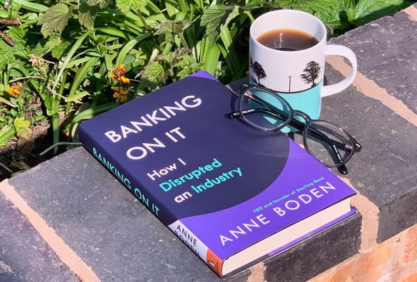 The book, Banking On It, by Anne Boden, on a garden wall with a cup of coffee and a pair of glasses